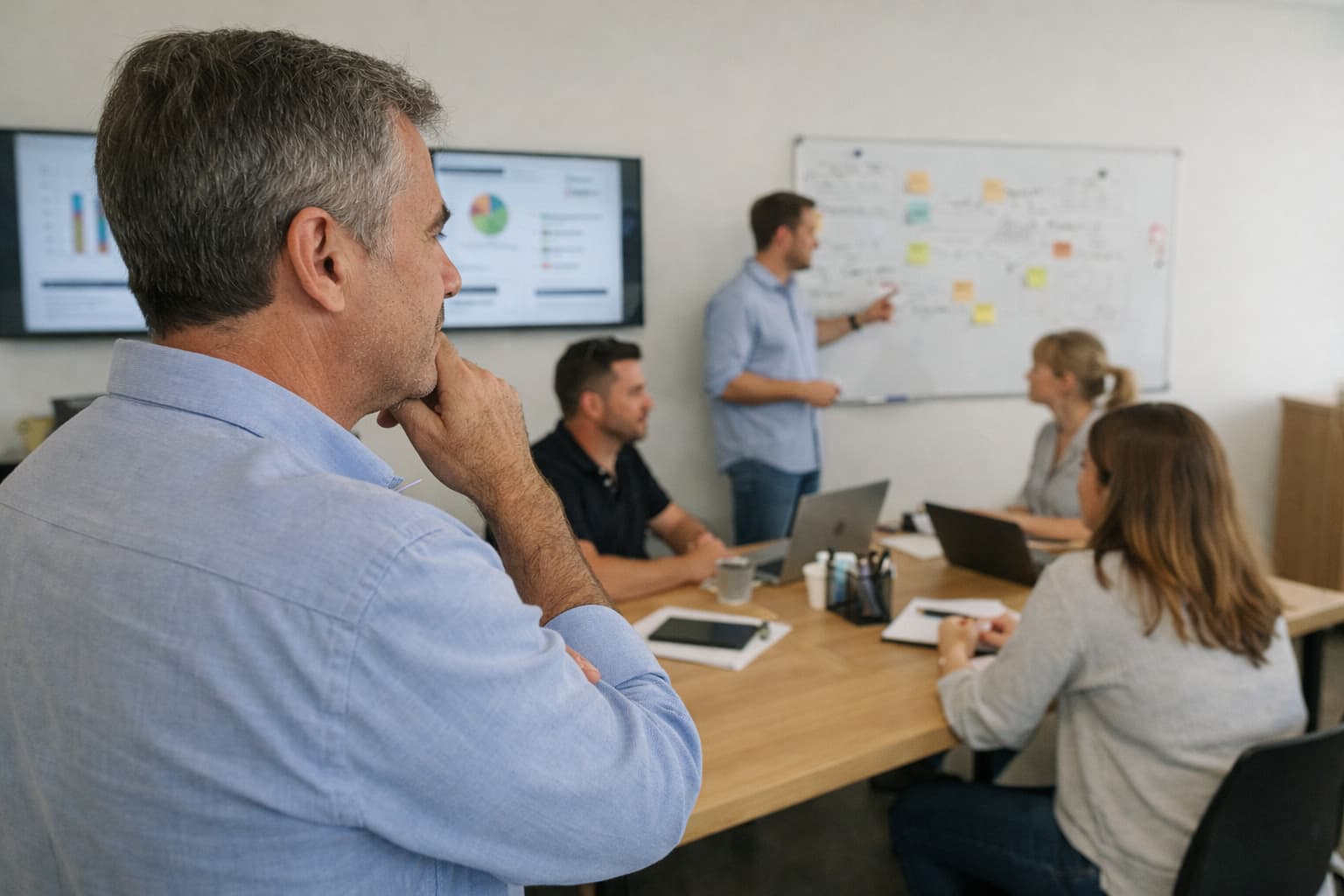 Empresário em pé observa equipe reunida em sala de reunião enquanto gestor conduz apresentação com indicadores no quadro.